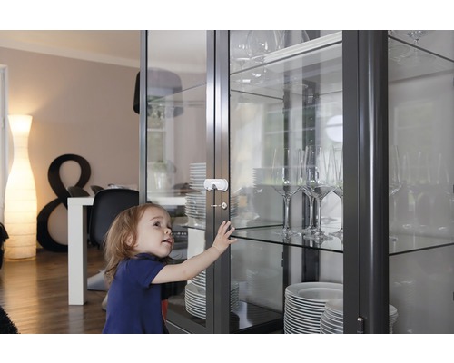 Un enfant essaie d''ouvrir une vitrine avec une sécurité enfant. Des assiettes et des verres se trouvent dans le meuble.