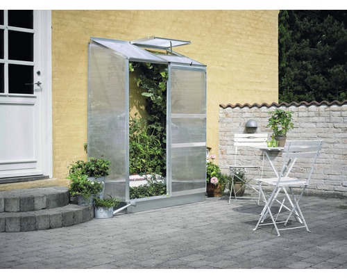 Anlehngewächshaus Halls Altan 2 Hohlkammerplatten 4 mm 132 x 69 cm Aluminium Gewächshaus aus Polycarbonat an einer Hauswand mit Pflanzen darin und Gartenmöbeln auf einer Terrasse.
