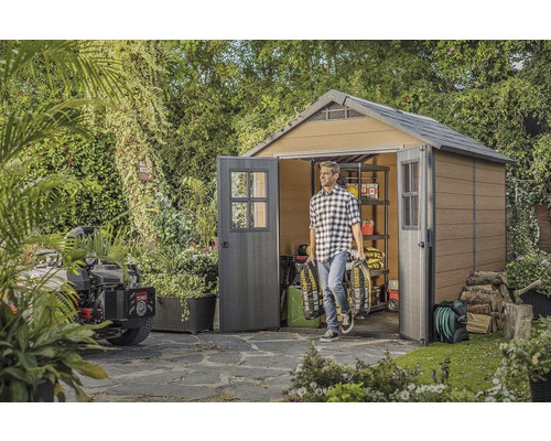 Ein Mann trägt Kohlesäcke aus einem Gartenhaus mit Regalen und Doppeltür in einem begrünten Garten.