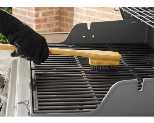 Eine Hand mit Grillhandschuh reinigt einen Grillrost mit einer Grillbürste mit Holzgriff.
