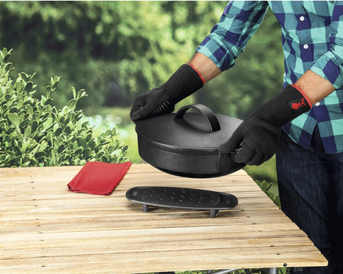 Personne avec des gants de barbecue manipule un four hollandais sur une table en bois à l''extérieur.