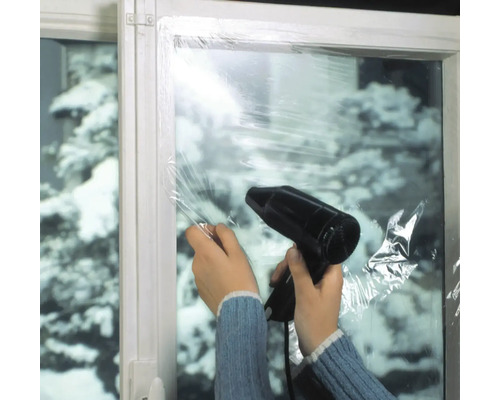 Person befestigt Fensterfolie mit einem Haartrockner an einem Fenster, um die Wärmeisolierung zu verbessern.