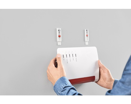 Une personne fixe une Fritzbox au mur avec deux clous adhésifs.