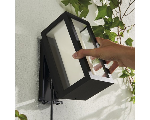 Une personne fixe une applique murale extérieure noire avec un verre transparent à un mur.