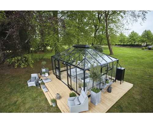Serre avec parois en verre et cadre noir sur une terrasse en bois dans le jardin, entourée d''arbres et d''espaces verts.