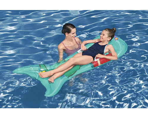 Une fille est allongée sur un matelas pneumatique en forme de poisson gonflable dans l''eau, à côté d''elle se tient une femme.