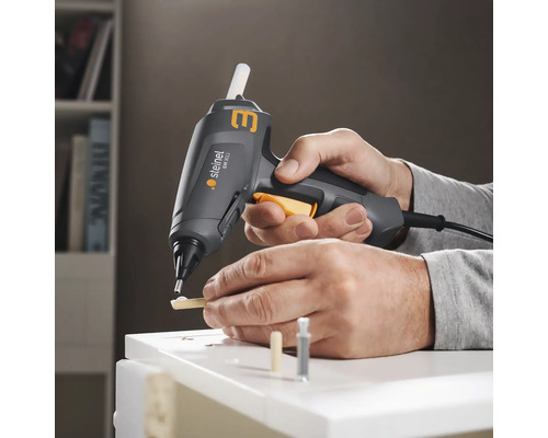 Une personne utilise un pistolet à colle Steinel pour coller des morceaux de bois sur une surface blanche.