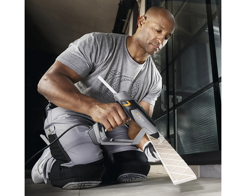 Un homme utilise un pistolet à colle chaude pour fixer un carreau. Le pistolet à colle est gris et noir. Le carreau est beige.